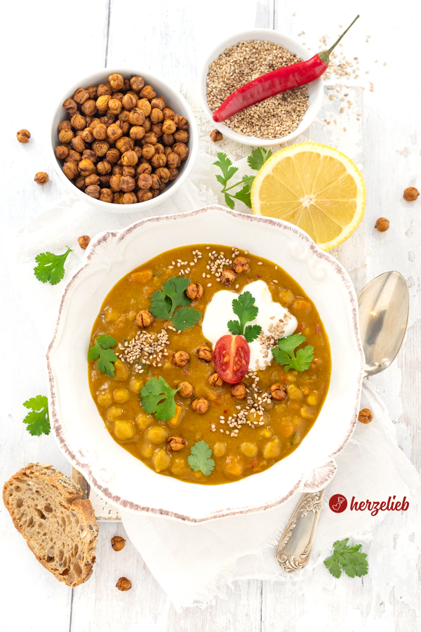 Eine weiße Schale mit Suppe nach einem Kichererbsensuppe Rezept von herzelieb. Angerichtet mit Brot, mit Koriander, einem Tomatenviertel und Joghurt. Von oben fotografiert. Im Hintergrund eine Schüssel mit Kichererbsen, eine Zitrone, Sesam und eine Chilischote