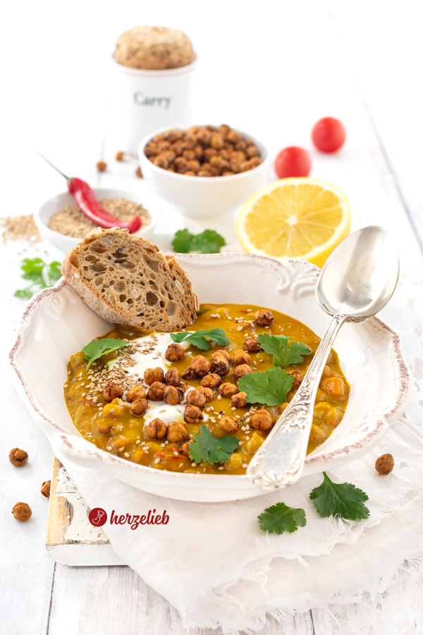 Ein Teller mit Suppe nach einem Kichererbsensuppe Rezept von herzelieb. Angerichtet mit Koriander, Joghurt und Brot. Auf dem Tellerrand liegt ein Silberlöffel. Im Hintergrunde eine Schale mit Sesam, in der eine rote Chili liegt, eine Schüssel mit Kichererbsen und eine Zitrone.