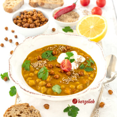 Leckere Kichererbsensuppe in einer weißen Schale. Angerichtet mit Joghurt, Koriander, einer Tomate und knusprigen Kichererbsen. Im Vordergrund ein Stück Brot. Im Hintergrund mehrere Brotscheiben, eine Schüssel mit Kichererbsen, eine Schale mit Sesam und einer roten Chilischote sowie eine Zitrone.