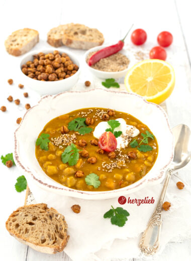 Leckere Kichererbsensuppe in einer weißen Schale. Angerichtet mit Joghurt, Koriander, einer Tomate und knusprigen Kichererbsen. Im Vordergrund ein Stück Brot. Im Hintergrund mehrere Brotscheiben, eine Schüssel mit Kichererbsen, eine Schale mit Sesam und einer roten Chilischote sowie eine Zitrone.