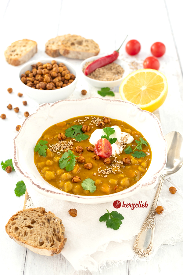 Leckere Kichererbsensuppe in einer weißen Schale. Angerichtet mit Joghurt, Koriander, einer Tomate und knusprigen Kichererbsen. Im Vordergrund ein Stück Brot. Im Hintergrund mehrere Brotscheiben, eine Schüssel mit Kichererbsen, eine Schale mit Sesam und einer roten Chilischote sowie eine Zitrone.