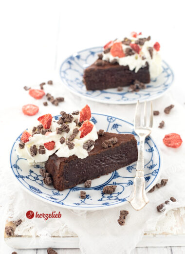 Ein Stück Schokoladenkuchen mit weichem Kern nach einem Kladdkaka Rezept aus Schweden von herzelieb auf einem kleinen Teller. Dekoriert mit Schokostücken und gefriergetrockneten Erdbeeren. Im Hitnergrund ein zweites Stück.