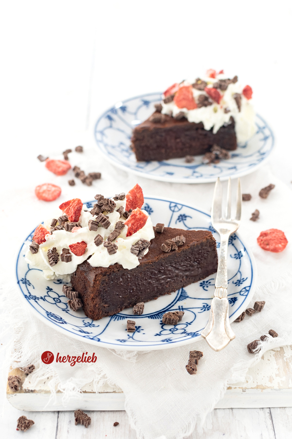 Ein Stück Schokoladenkuchen mit weichem Kern nach einem Kladdkaka Rezept aus Schweden von herzelieb auf einem kleinen Teller. Dekoriert  mit Schokostücken und gefriergetrockneten Erdbeeren. Im Hitnergrund ein zweites Stück.