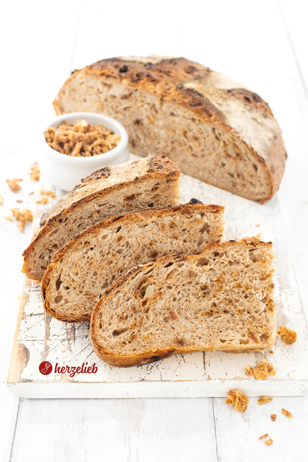 Ein angeschnittenes Vollkornbrot, das mit Kohl und Röstzwiebeln gebacken nach einem Kohlbrot Rezept von herzelieb gebacken ist. . Im Vordergrund lehnen 2. Scheiben von dem Brot vor einem Stück des kräfigen Brotes. Dahinter eine Schale mit Röstzwiebeln. Hinten rechts ein halber Brotlaib.