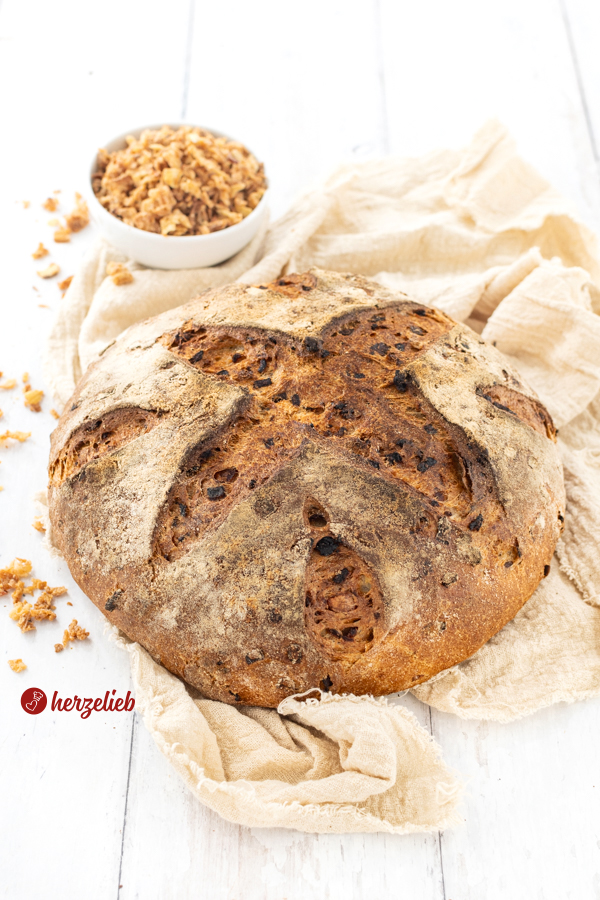 Ein Kohlbrot mit Röstzwiebeln nach einem Vollkornbrot Rezept von herzelieb. Der Brotlaib ist auf einem beigefarbenen Tuch platziert, mit Mehl bestäubt und mehrfahc eingeschnitten. Im Hintergrund steht ein Schälchen mit Röstzwiebeln. 