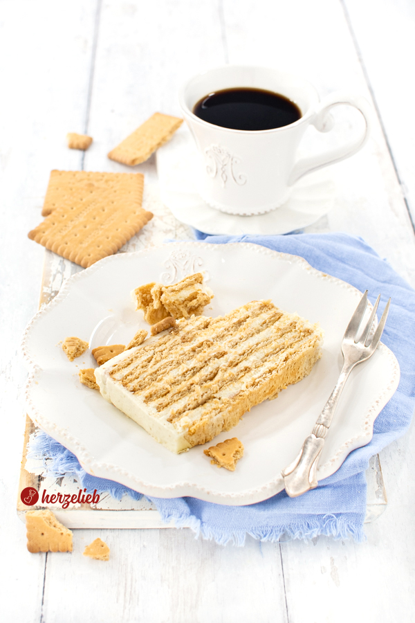 Ein Stück Kekskuchen nach einem Kondensmilichkuchen Rezept ohne backen von herzelieb. Auf dem Tellerrand liegt eine Kuchengabel. im Hintergrund eine Tasse Kaffee, links daneben einige Kekse.