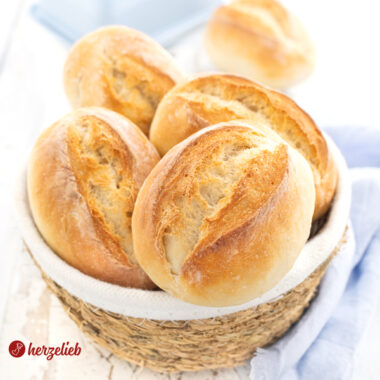 Ein kleines Körbchen mit goldbraun gebackenen Brötchen nach einem Frühstücksbrötchen Rezept von herzelieb. Im Hintergrund steht eine blaue Butterglocke. Um den Korb herum liegt ein hellblaues Tuch.