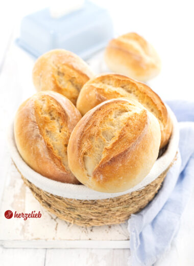 Ein kleines Körbchen mit goldbraun gebackenen Brötchen nach einem Frühstücksbrötchen Rezept von herzelieb. Im Hintergrund steht eine blaue Butterglocke. Um den Korb herum liegt ein hellblaues Tuch.