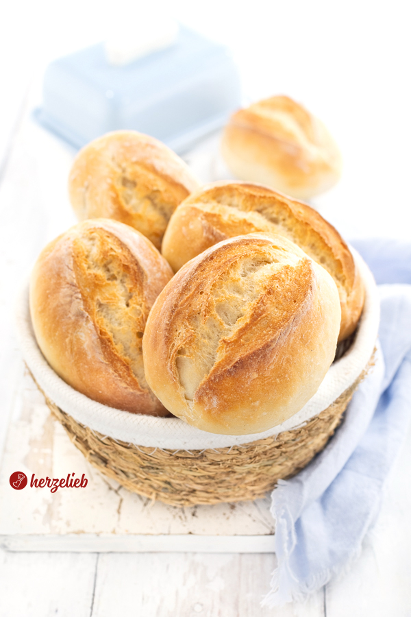 Ein kleines Körbchen mit goldbraun gebackenen Brötchen nach einem Frühstücksbrötchen Rezept von herzelieb. Im Hintergrund steht eine blaue Butterglocke. Um den Korb herum liegt ein hellblaues Tuch.