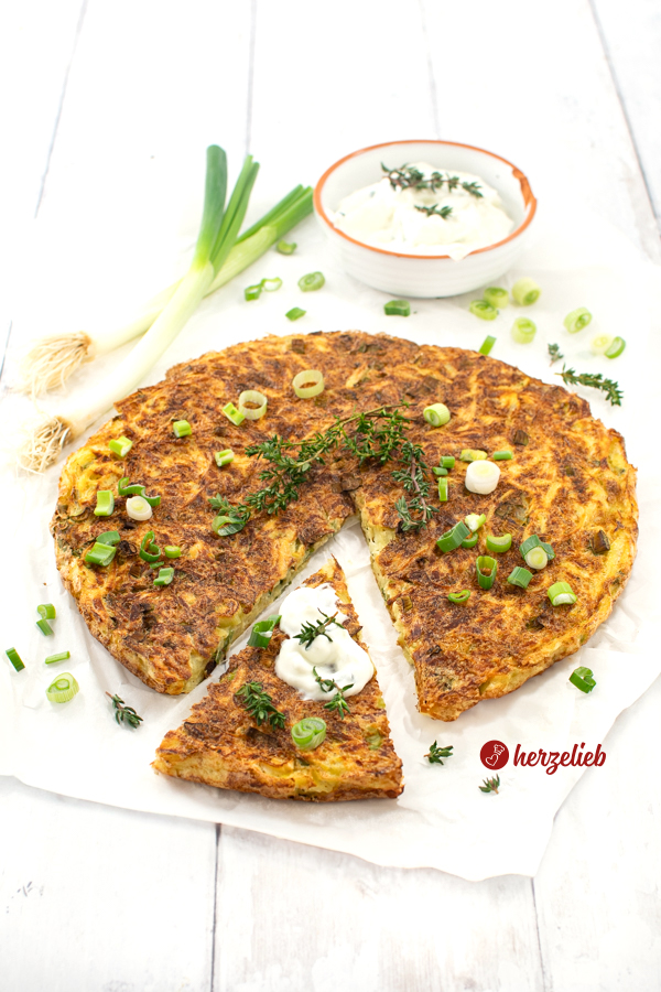 Eine angeschnittene, knusprige Kartoffeltarte nach einem Kartoffelkuchen Rezept von herzelieb auf Backpapier. Dekoriert mit Thymian und Lauchzwiebel Röllchen. Im Hintergrund ein Schälchen Tzatziki, zwei Gewürzgefäße und zwei Lauchzwiebeln.