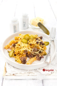 Ein weißer, flacher Teller mit hohem Rand mit einem Auflauf nach einem Burger-Nudelauflauf Rezept von herzelieb. Man sieht das hacfklich Nudeln, Gurkenstücke und den Käse. Im Hintergrund ein Pfeffer und ein Salzstreuer.