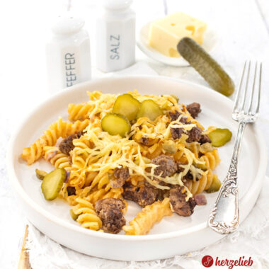 Ein weißer, flacher Teller mit hohem Rand mit einem Auflauf nach einem Burger-Nudelauflauf Rezept von herzelieb. Man sieht das hacfklich Nudeln, Gurkenstücke und den Käse. Im Hintergrund ein Pfeffer und ein Salzstreuer.