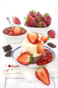 Ein helles Törtchen mit einem dunklem Boden nach einem Grießtörtchen Rezept von herzelieb. Dekoriert mit Erdbeergrütze und halben Erdbeeren.Im Hintergrund ein Schälchen mit roter Grütze und einer Schale Erdbeeren. Neben, vor und hinter dem Teller mit dem Törtchen lieben Schokoladenstücke und halbe sowie ganze Erdbeeren.