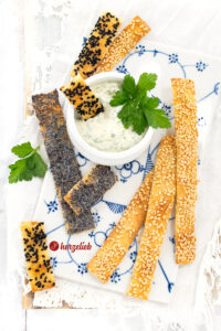 Einige Brotstangen nach einem Potatiskex Rezept aus Schweden von herzelieb. Angerichtet auf einem Brett, mit einem Dip, in dem ein Stück Brotstick und ein Petersilienblatt steckt. Die Stangen sind bestreut mit schwarzem Sesam, geröstetem Sesam und Mohn.