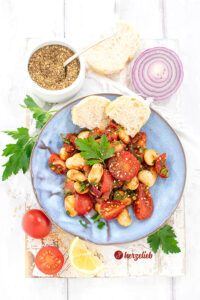 Auf einem blauen Teller ist ein Salat mit Tomaten und weißen Bohnen nach einem Tomatensalat Rezept mit Riesenbohnen von herzelieb angerichtet. Dekoriert mit Petersilie, bestreut mit Zatar und serviert mit Baguettebrot.