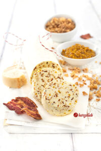 Eine angeschnittene Rolle Gewürzbutter nach einem Zwiebel-Bacon-Butter Rezept von herzelieb. Vor der Rolle liegen zwei dicke Scheiben von der Butter. Vorne rechts ein Stück knuspriger Bacon.