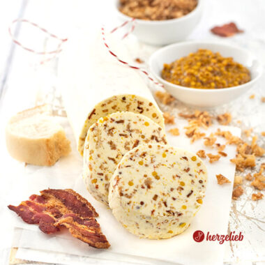 Eine angeschnittene Rolle Gewürzbutter nach einem Zwiebel-Bacon-Butter Rezept von herzelieb. Vor der Rolle liegen zwei dicke Scheiben von der Butter. Vorne rechts ein Stück knuspriger Bacon.