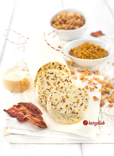 Eine angeschnittene Rolle Gewürzbutter nach einem Zwiebel-Bacon-Butter Rezept von herzelieb. Vor der Rolle liegen zwei dicke Scheiben von der Butter. Vorne rechts ein Stück knuspriger Bacon.