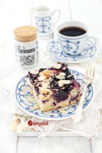Ein Stück Blaubeer-Butterkuchen nach einem Blechkuchen Rezept von herzelieb. Angerichtet mit einer Kuchengabel auf einem blauweißen Teller. Im Hintergrund eine Tasse Kaffee, ein Gewürzgefäß und ein Milchkännchen.