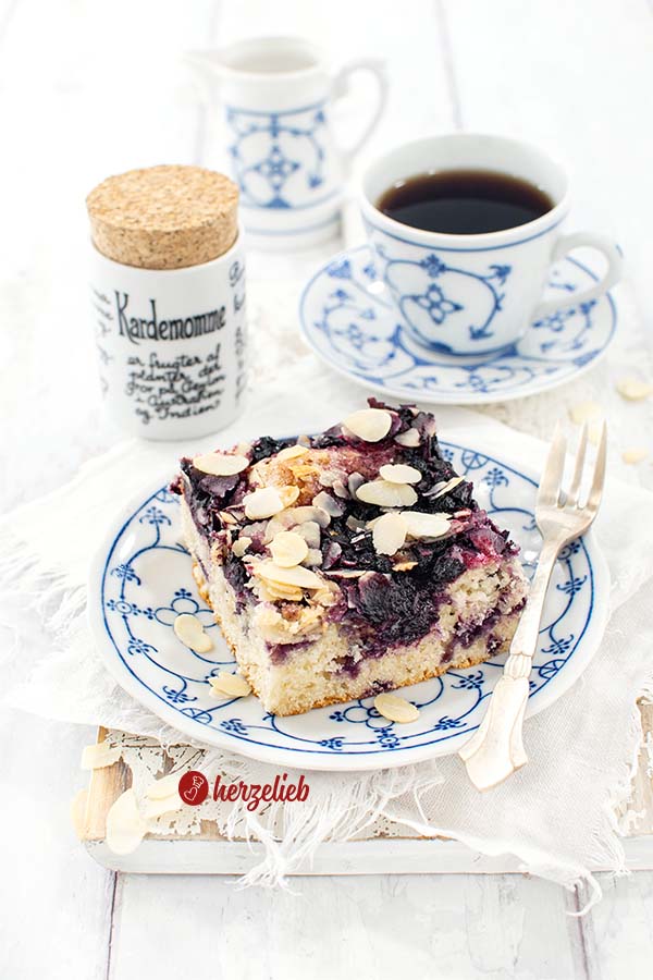 Ein Stück Blaubeer-Butterkuchen nach einem Blechkuchen Rezept von herzelieb. Angerichtet mit einer Kuchengabel auf einem blauweißen Teller. Im Hintergrund eine Tasse Kaffee, ein Gewürzgefäß und ein Milchkännchen.