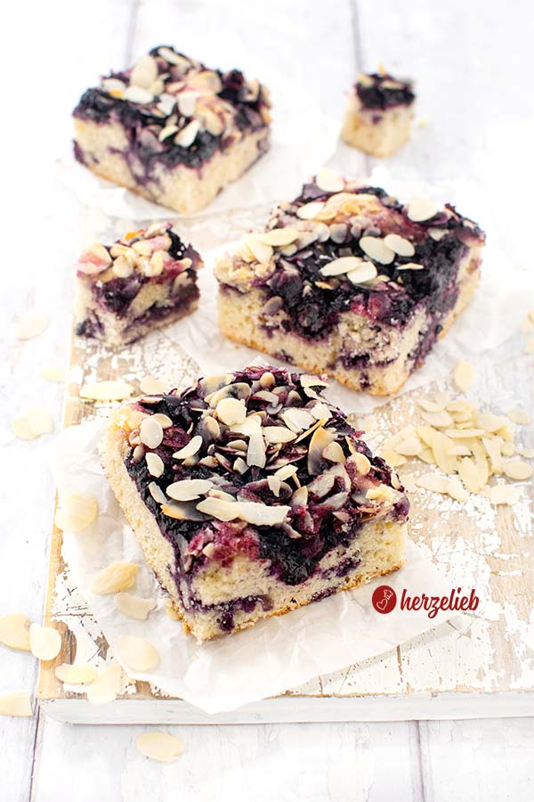 3 Stücke Blaubeer-Butterkuchen auf einem weißen Holzbrett. Die quadratischen Stücke sind mit Mandelblättchen bestreut. 