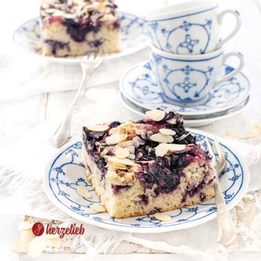Auf einem weiß-blau gemustertem Teller ein Stück Kuchen nach ienem Blaubeer-Butterkuchen Rezept von herzelieb. Bstreut mit Mandelblättchen. Im Hintergrund zwei ineinander gestapelte Tassen und ganz hinten ein weiteres Stück Kuchen