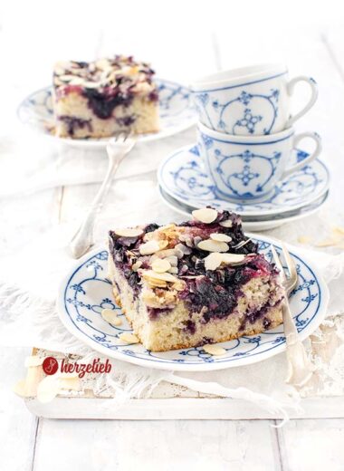Auf einem weiß-blau gemustertem Teller ein Stück Kuchen nach ienem Blaubeer-Butterkuchen Rezept von herzelieb. Bstreut mit Mandelblättchen. Im Hintergrund zwei ineinander gestapelte Tassen und ganz hinten ein weiteres Stück Kuchen