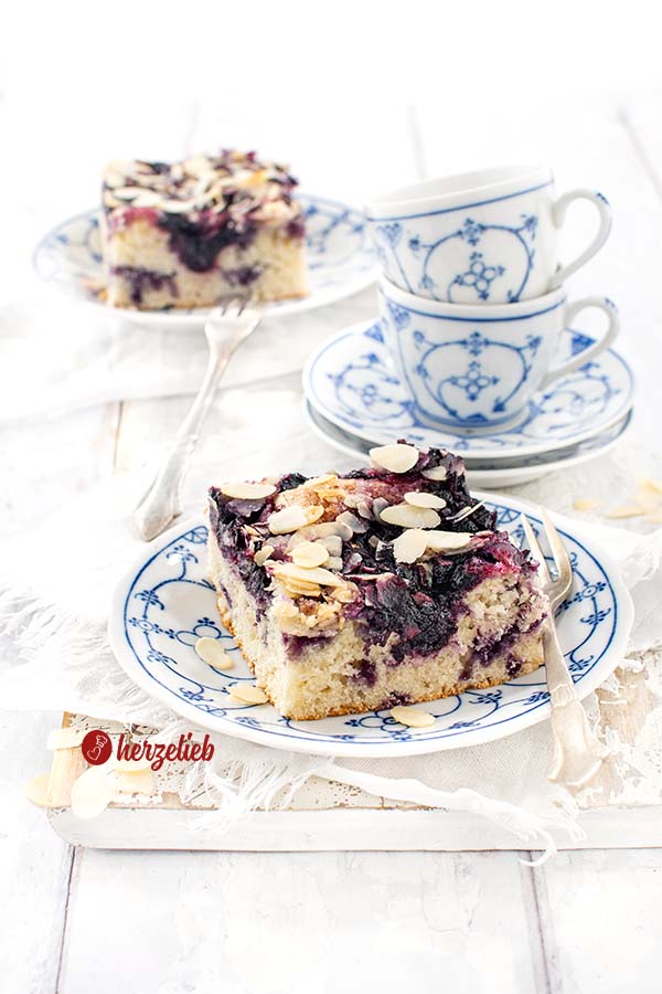 Auf einem weiß-blau gemustertem Teller ein Stück Kuchen nach ienem Blaubeer-Butterkuchen Rezept von herzelieb. Bstreut mit Mandelblättchen. Im Hintergrund zwei ineinander gestapelte Tassen und ganz hinten ein weiteres Stück Kuchen 