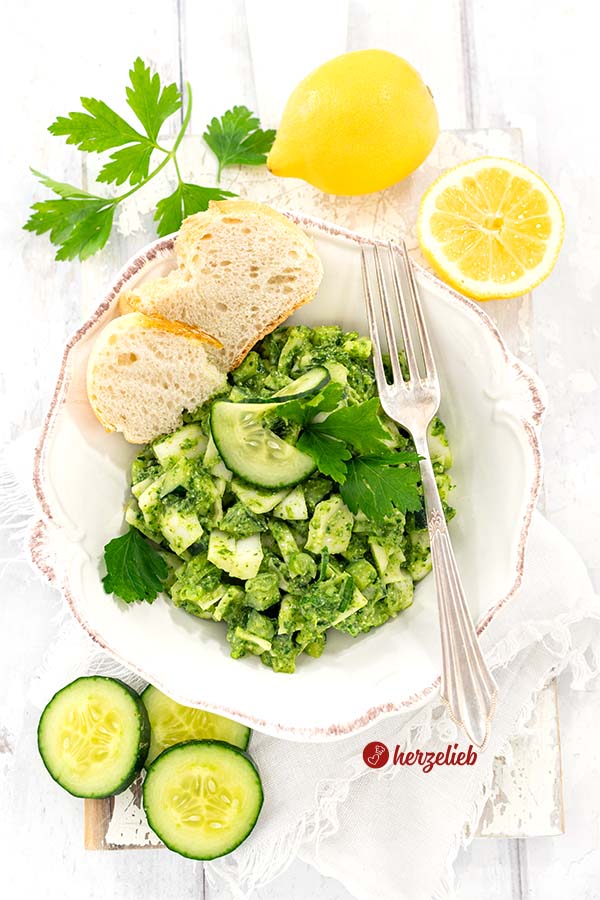 Ein Schale mit einem Kohlsalat oder Gurkensalat nach einem Grüne Göttin Salat Rezept von herzelieb. Angerichtet mit Brotscheiben, Gurkenscheiben und Petersilie. Auf dem Schüsselrand liegt eine Gabel. Im Hintergrund eine halbe und eine ganze Zitrone