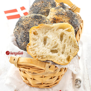 Fluffige Mohnebrötchen nach einem Håndværkere Rezept aus Dänemark in einem Körbchen. Obenauf liegt ein halbiertes Brötchen. Im Hintergrund und im Vordergrund je ein Dannebrog.