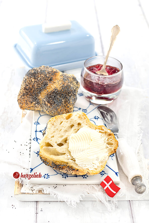Ein blauweißes Frühstücksbrett aus Porzellan mit einer Mohnbrötchen-Hälfte, die mit Butter bestrichen ist. Im Hintergrund ein weiteres Håndværkere Brötchen nach einem Rezept aus Dänemark.