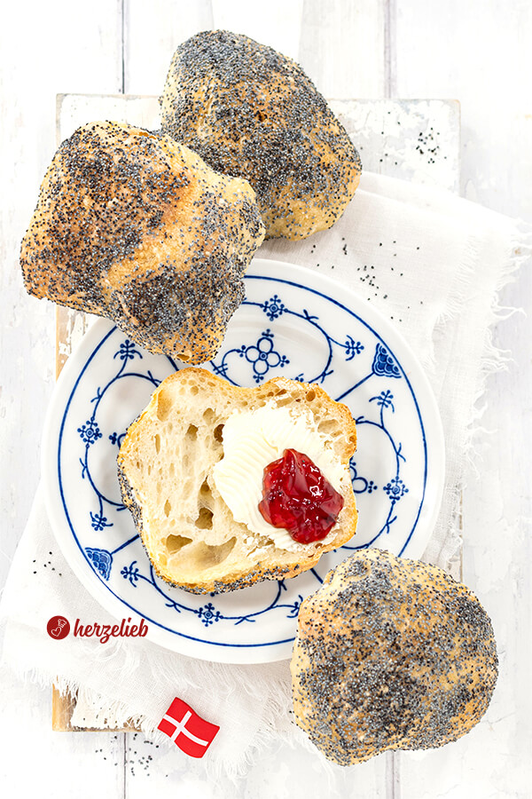 Ein weißblauer Teller mit einem mit Butter und Marmelade bestrichenen Brötchen nach einem Håndværkere Rezept aus Dänemark von herzelieb. Rundherum 3 weitere Mohnbrötchen davon. 