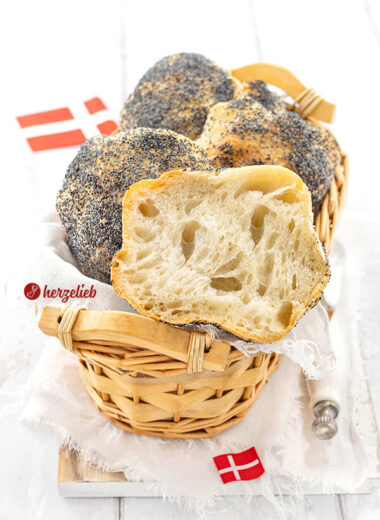 Fluffige Mohnebrötchen nach einem Håndværkere Rezept aus Dänemark in einem Körbchen. Obenauf liegt ein halbiertes Brötchen. Im Hintergrund und im Vordergrund je ein Dannebrog.