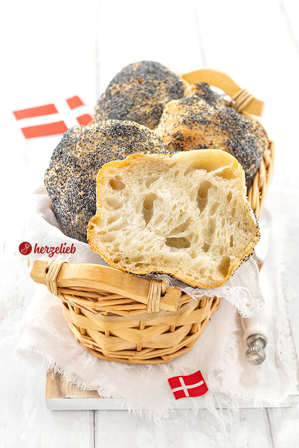 Fluffige Mohnbrötchen nach einem Håndværkere Rezept aus Dänemark in einem Körbchen. Obenauf liegt ein halbiertes Brötchen. Im Hintergrund und im Vordergrund je ein Dannebrog.