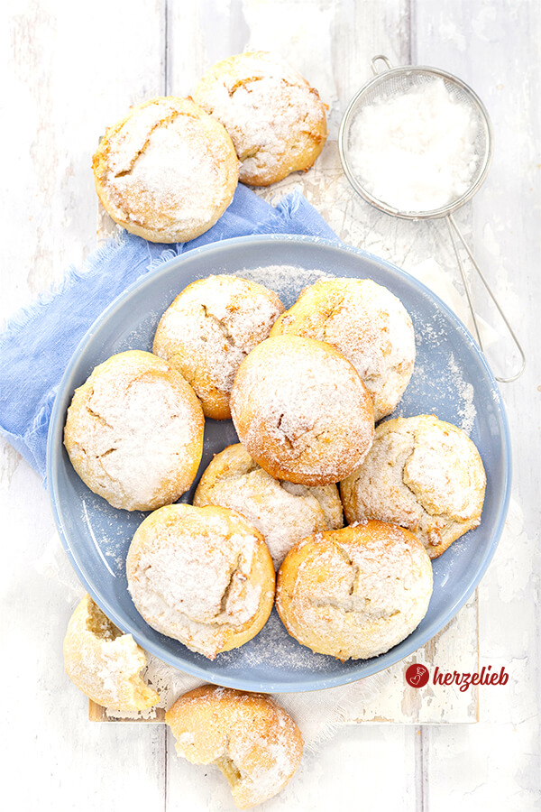 Goldgelbe Plätzchen nach einem Käsekuchen Kekse Rezept von herzelieb mit Puderzucker bestäubt, auf einem blauen Teller und einem blauen Tuch, daneben ein Sieb mit Puderzucker und ein halb gegessener Keks auf hellem Untergrund.