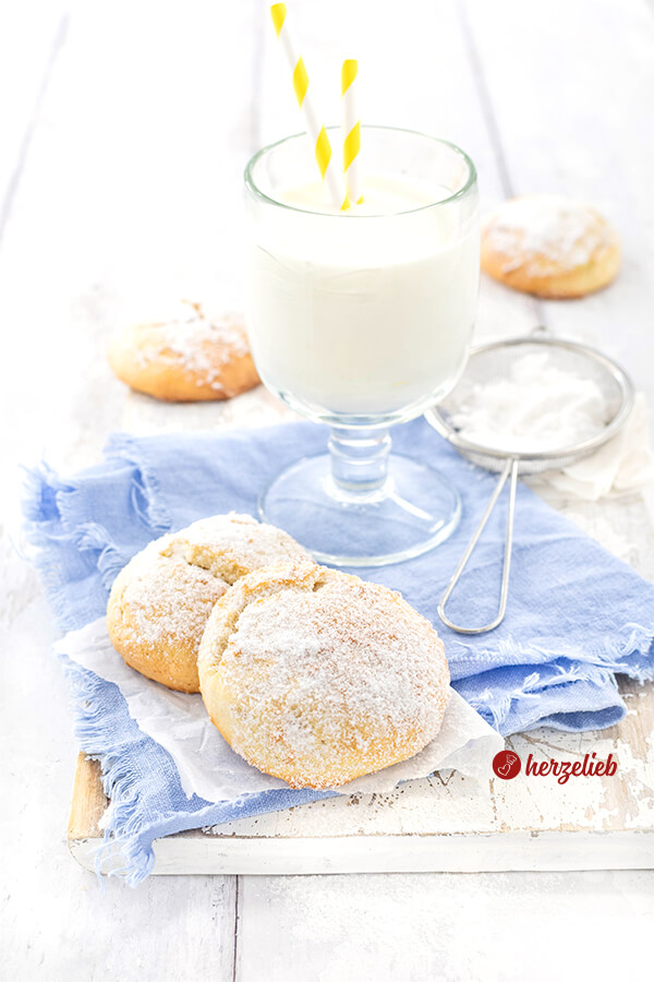 Zwei mit Puderzucker bestäubte Plätzchen nach einen Käsekuchen Kekse Rezept von herzelieb auf blauem Tuch, daneben ein Glas Milch mit zwei gelb-weißen Strohhalmen und ein Sieb mit Puderzucker.