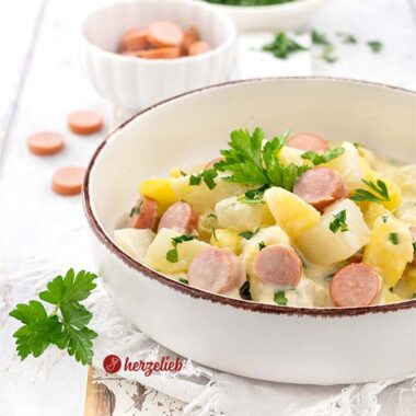Eine Keramikschale mit einem dunklem Rand, gefüllt mit einen Kartoffel-Kohlrabi-Topf. Im Eintopf sieht man Würstchen, Kartoffelstücke, Kohlrabi-Würfel. Dekoriert mit Petersilie.