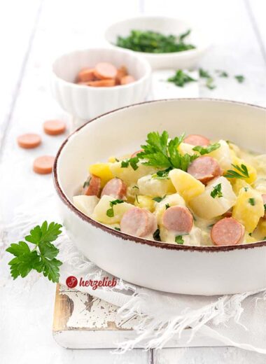 Eine Keramikschale mit einem dunklem Rand, gefüllt mit einen Kartoffel-Kohlrabi-Topf. Im Eintopf sieht man Würstchen, Kartoffelstücke, Kohlrabi-Würfel. Dekoriert mit Petersilie.