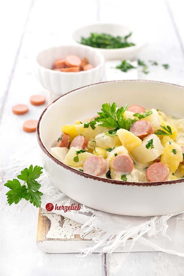 Eine Keramikschale mit einem dunklem Rand, gefüllt mit einen Kartoffel-Kohlrabi-Topf. Im Eintopf sieht man Würstchen, Kartoffelstücke, Kohlrabi-Würfel. Dekoriert mit Petersilie.