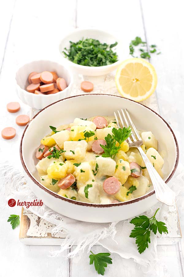 Eine Schale mit einem Kohlrabigericht nach einem Kartoffel-Kohlrabi-Topf Rezept von herzelieb. Angerichtet mit Würstchen in einer weißen Soße, dekoriert mit Petersllie. Im Hintergrund eine Zitron, eine Schale mit Würstchenscheiben und eine Schälchen gehackte Petersilie. 
