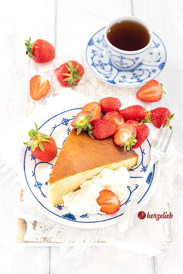 Milchmädchenkuchen mit Sahne und frischen Erdbeeren auf einem blauweißem Teller. Auf dem Tellerrand eine silberne Kuchengabel. Im Hintergrund eine Tasse Tee und weitere frische Früchte