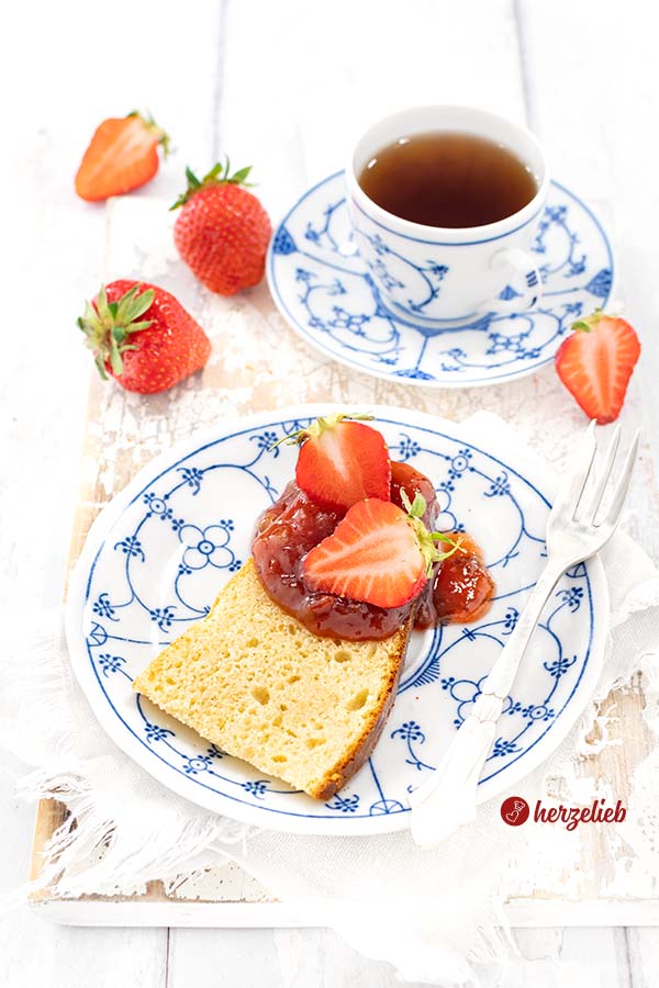 Ein Stück goldgelb gebackener Kuchen nach einem Milchmädchenkuchen Rezept von herzelieb. Angerichtet auf einem blauweißen Teller mit Erdbeer-Rhabarber-Marmelade und frischen Erdbeeren von herzelieb. Im Hintergrund eine Tasse Tee und weitere Erdbeeren. 