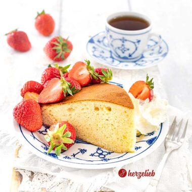 Ein Stück Milchmädchenkuchen auf einem blauweißen Teller. Angerichtet mit Sahne nd frischen Erdbeeren. . Im Hintergrund weitere frische Früchte und eine Tasse Tee