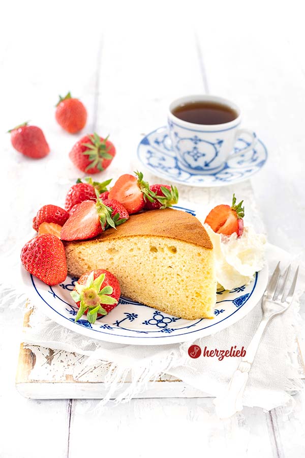 Ein Stück Milchmädchenkuchen auf einem blauweißen Teller. Angerichtet mit Sahne nd frischen Erdbeeren. . Im Hintergrund weitere frische Früchte und eine Tasse Tee