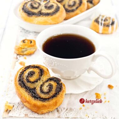 Eine Tasse Kaffee mit einer goldbraun gebackene Gebäckschnecke nach einem Mohnschnecken Rezept von herzelieb. Im Hintergrund eine ovale Porzellanplatte mit weiteren Hefeschnecken.