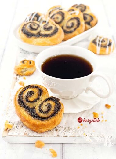Eine Tasse Kaffee mit einer goldbraun gebackene Gebäckschnecke nach einem Mohnschnecken Rezept von herzelieb. Im Hintergrund eine ovale Porzellanplatte mit weiteren Hefeschnecken.