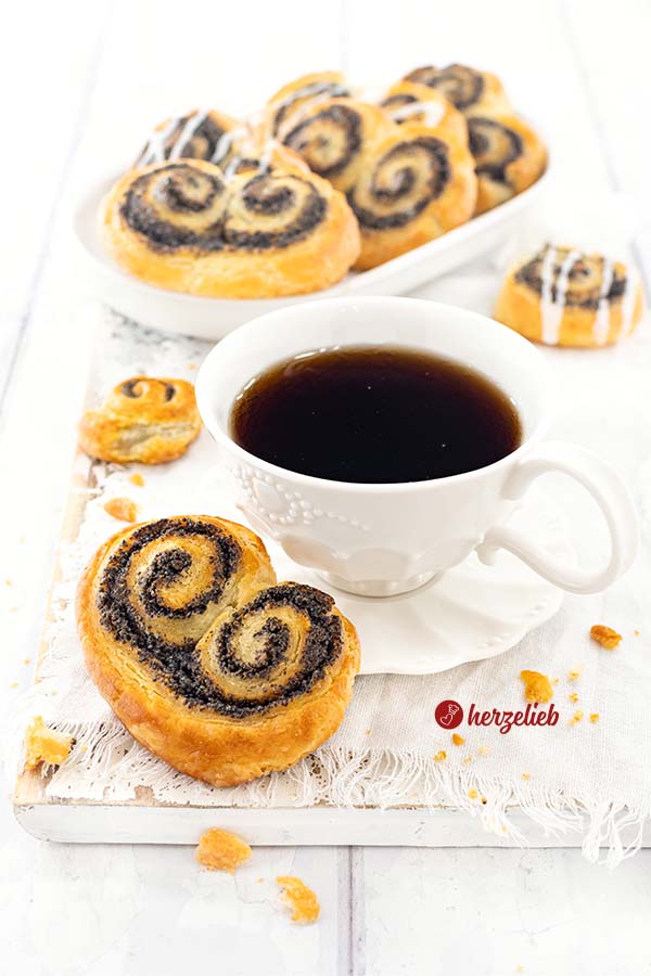 Eine Tasse Kaffee mit einer goldbraun gebackene Gebäckschnecke nach einem Mohnschnecken Rezept von herzelieb. Im Hintergrund eine ovale Porzellanplatte mit weiteren Schnecken.