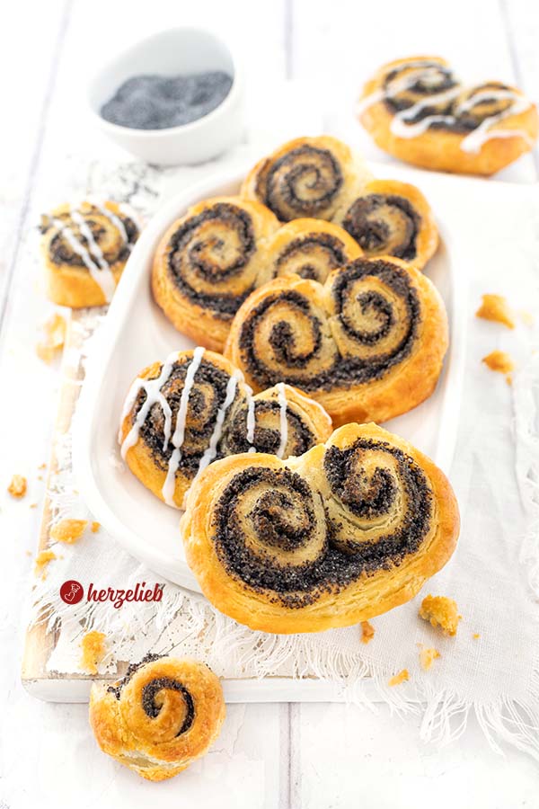 5 Mohnschnecken auf einer weißen, ovalen Porzellanplatte. Im Hintergrund eine weitere Gebäckschnecke und eine Schale mit Mohn.
