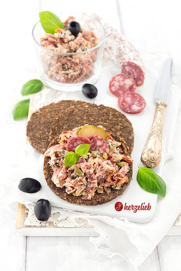 3 Scheiben Schwarzbrot, die übereinanderliegen. Das oberste ist mit dem Rustami-Aufstrich nach einem Salami-Aufstrich Rezept von herzelieb bestrichen. Im Hintergrund eine angeschnittene Salami, ein Messer und ein Gläschen mit dem Brotaufstrich. Dekoriert mit Oliven, Salamischeiben und Basilikum.