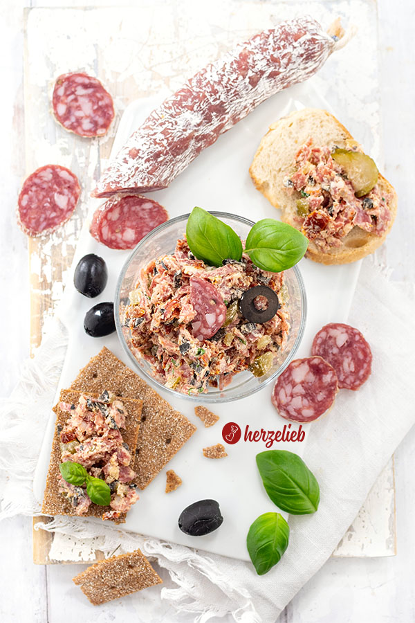 In einem Glas sieht man einen Rustami-Aufstrich – der Salami-Brotaufstrich ist dekoriert mit Salamischeiben, Olive, Basilikum. Vorne links 2 Knäckebrote mit dem Brotaufstrich. Hinten rechts ein Stück Baguette damit. Hinten links eine angeschnittene Salami.